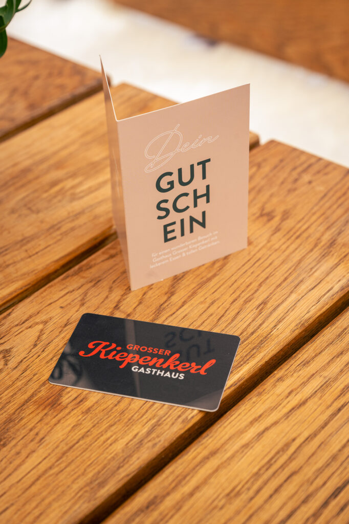 Schwarze Gutscheinkarte im Scheckkartenformat mit dem Logo des Grossen Kiepenkerls versehen (Farben rot/weiss), daneben Gutscheinhülle in Beige zum verpacken