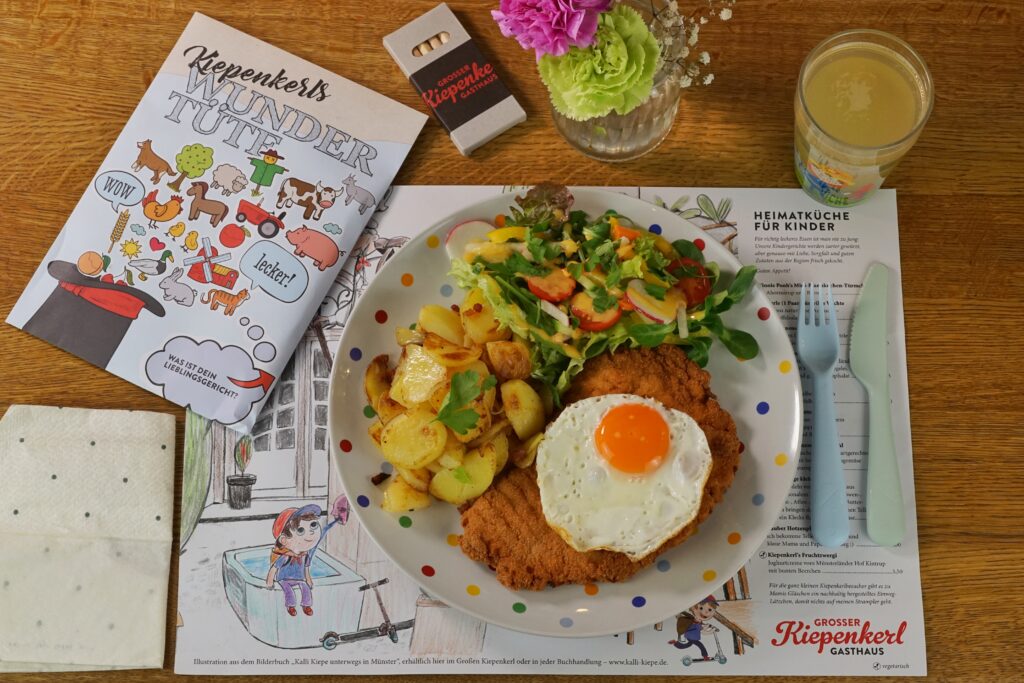 Kinderessen, auf weißem Teller mit bunten Punkten angerichtetes Schnitzel mit Spiegelei, dazu Bratkartoffeln und Salat.