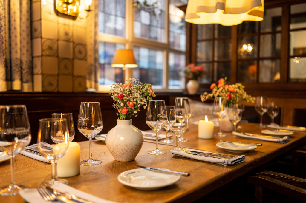 Eingedeckter Tisch im Gasthaus, mit Vorspeisentellern, Stoffservietten, Besteck, Wasser- und Weingläsern, dekoriert mit orange/weißen Blumen in einer hellen Vase.