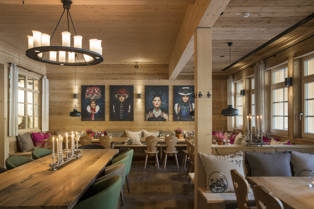 Stilvoller Gastraum im Alpenstil mit Holzwänden, großen Fenstern und warmem Kerzenlicht. An der Wand hängen moderne Porträts in traditioneller Tracht, die dem Raum eine besondere Atmosphäre verleihen.