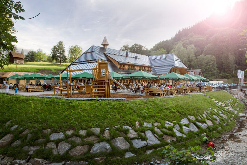 Großer Biergarten mit Holzhaus im traditionellen Stil, umgeben von Natur und Wald. Gäste sitzen unter grünen Sonnenschirmen an Holzbänken, im Vordergrund ein Spielplatz und ein Bachlauf mit spielenden Kindern