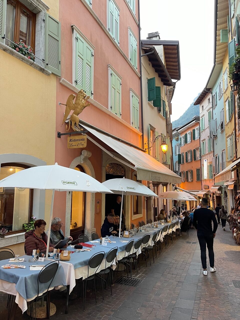 Eine kleine gemütliche Gasse, mit pastellfarbenden Häusern. Auf der linken Seite befindet sich das Restaurant Leon d’Oro. Vor der Fassade sind einige Außenplätze, die teils mit einer Markise überdacht sind, für die anderen Plätze gibt es weiße Schirme.