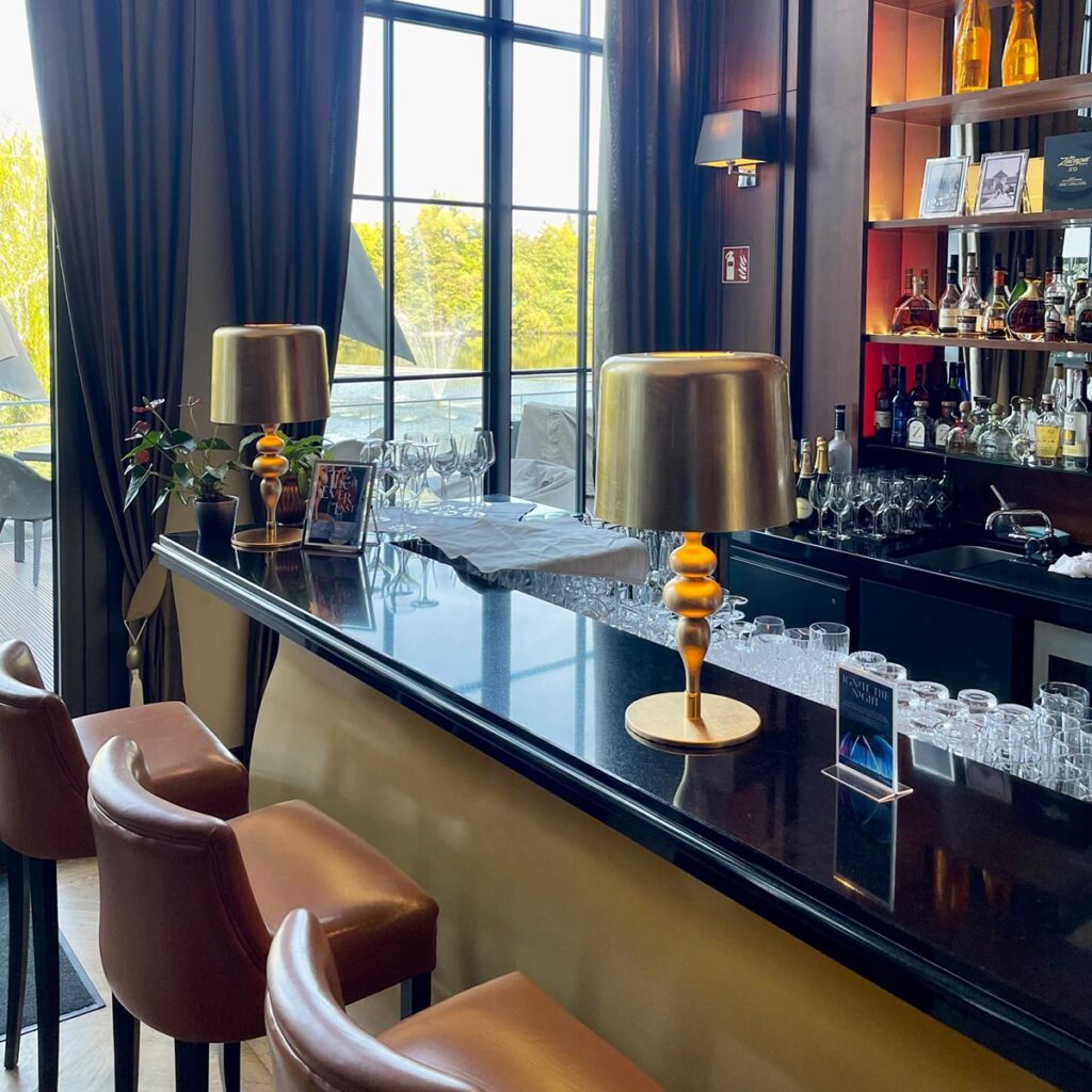 Dunkel glänzende Theke des Restaurants Leon d’Oro, mit goldenen Tischlampen und stilvollen braunen Ledersesseln. Im Hintergrund große Fenster, mit Blick ins Grüne und ein Regal mit edlen Spirituosenflaschen.
