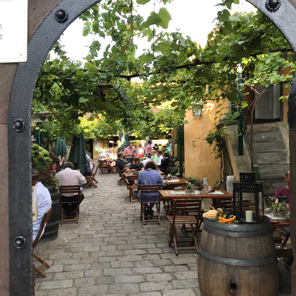 Gemütlicher Innenhof, geziert mit Weinreben. Tische und Stühle aus Holz, dunkelgrüne Sonnenschirme und mit Blumen dekorierte Weinfässer. Wirft man einen Blick in den Hof, wird dieser durch einen hölzernen Bogen geziert.