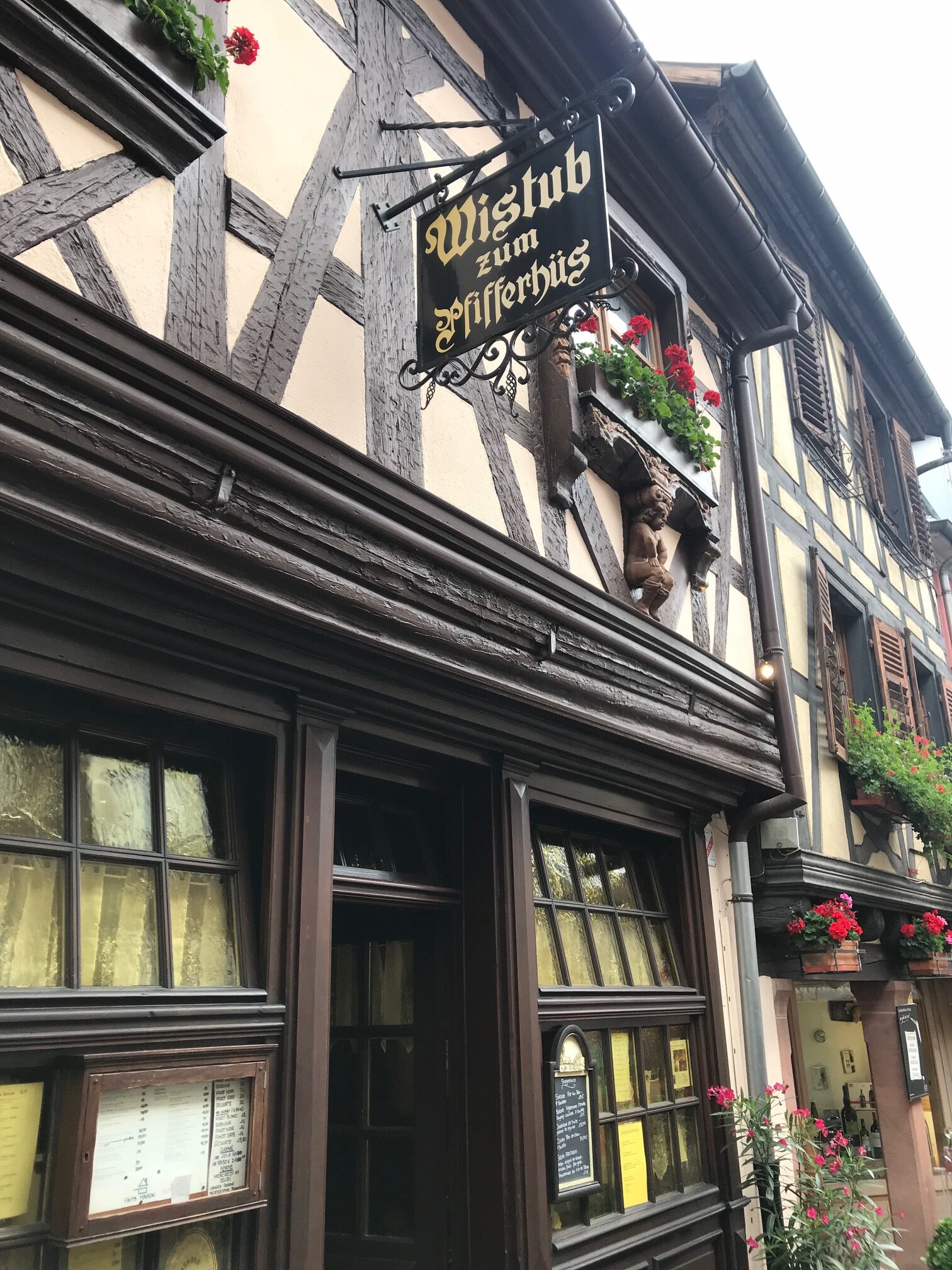 Fachwerkhaus mit dunkler Holzfassade und dem Wirtshausschild „Wistub zum Pfifferhüs“; Fenster mit weißen Vorhängen, Blumenkästen mit roten Geranien und Holzfigur an der Hauswand