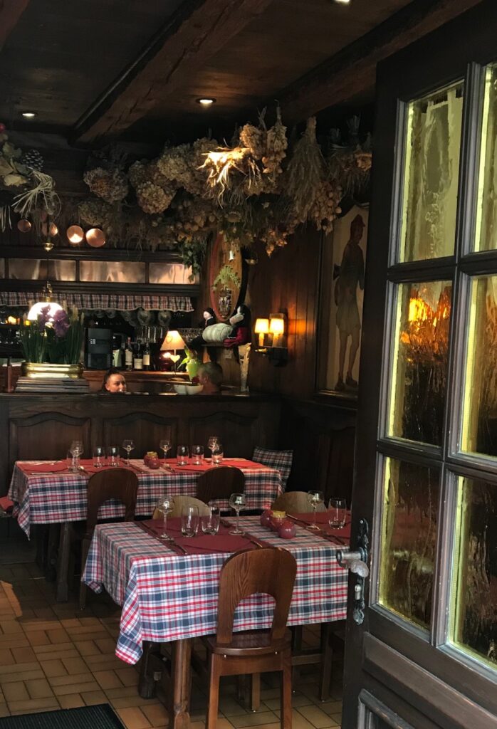 Urig-gemütlicher Gastraum mit dunkler Holzvertäfelung, Holzdecke mit herabhängenden Trockenblumen und rustikalen Stühlen; Tische mit blau-rot-weiß karierten Tischdecken liebevoll eingedeckt.