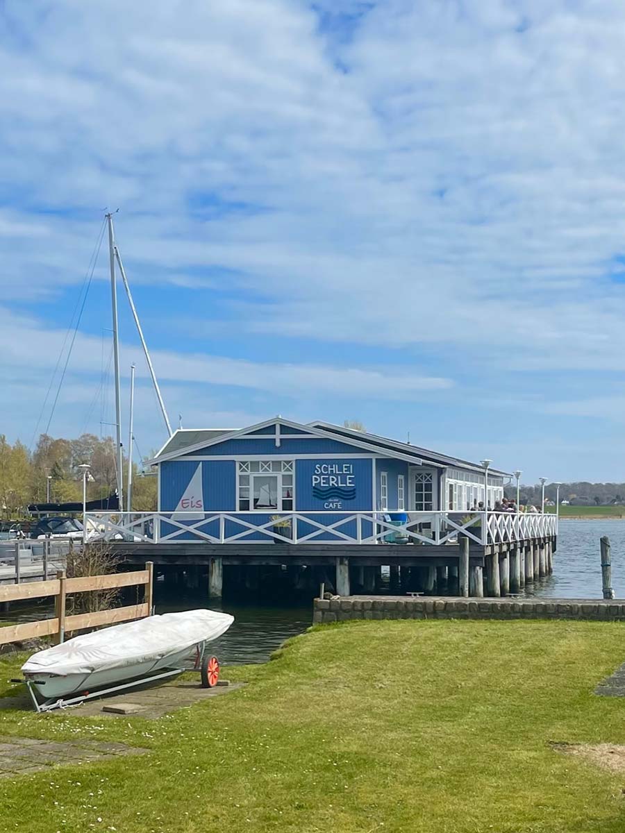 Blau-weißes Café, mit der Aufschrift „Schleiperle – Café“, auf Pfählen im Wasser, mit umlaufender Holzterrasse; davor ein kleines, abgedecktes Boot an Land; im Hintergrund Segelboote und Uferlandschaft.
