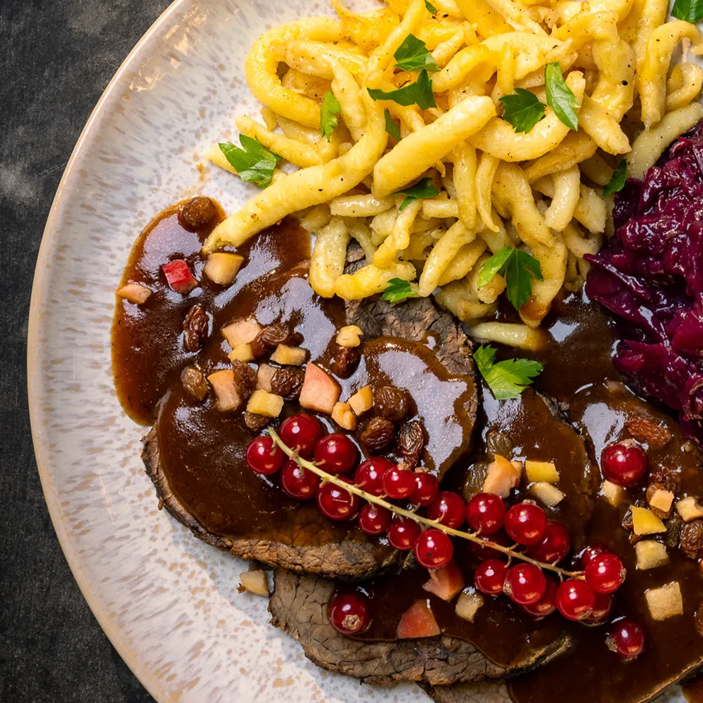 Ausschnitt eines schön angerichteten Tellers mit Sauerbraten, Rotkohl und Spätzle, dekoriert mit Johannisbeeren und frischen Kräutern.