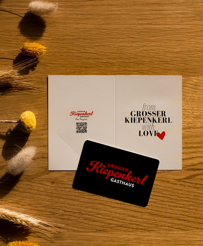 mittig: Gutschein vom Gasthaus Grosser Kiepenkerl (Umschlag & Karte), auf der linken Seite: als Deko gelb- & beigefarbene Trockenblumen, auf einem Holztisch liegend